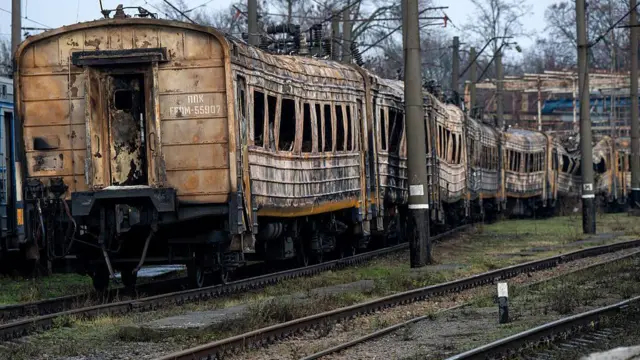 Довгий ряд сильно пошкоджених і обгорілих вагонів електрички, що стоять на залізничних коліях. Корпуси вагонів почорнілі, з вибитими або деформованими вікнами, видно сліди пожежі та руйнувань по всій довжині складу.