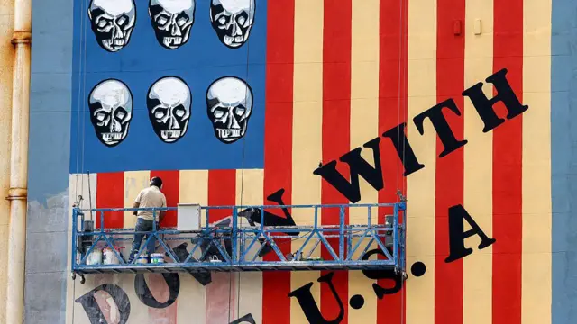 A man on a platform applies a fresh coat of paint to the landmark anti-US mural drawn on the wall of a building on Karim Khan Zand Avenue in Tehran on April 8, 2025 with the slogan "Down with the USA" and skulls replacing the stars on the US flag.