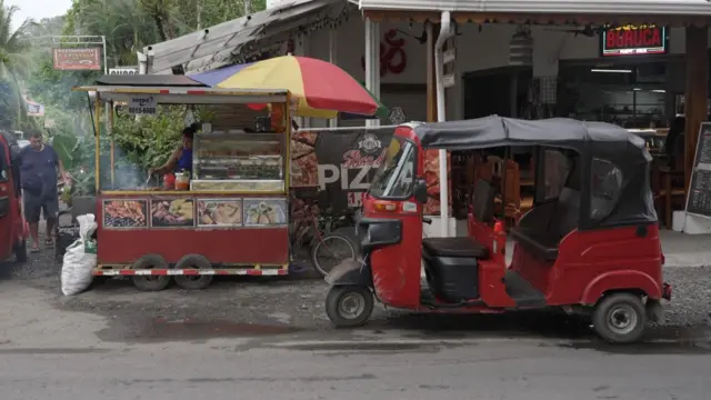 Un vehículo tuk tuk rojo está estacionado a un lado de la vía principal de Puerto Viejo mientras una mujer atiende un puesto de comida rápida que ofrece comida tradicional del Caribe costarricense.