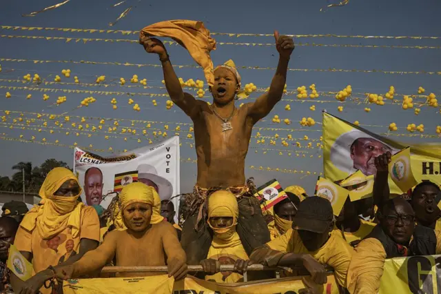 A group of pro-government supporters in yellow attire hold on to a railing. A man covered in yellow dye sits on the shoulders of another, waving a yellow cloth in the air. 