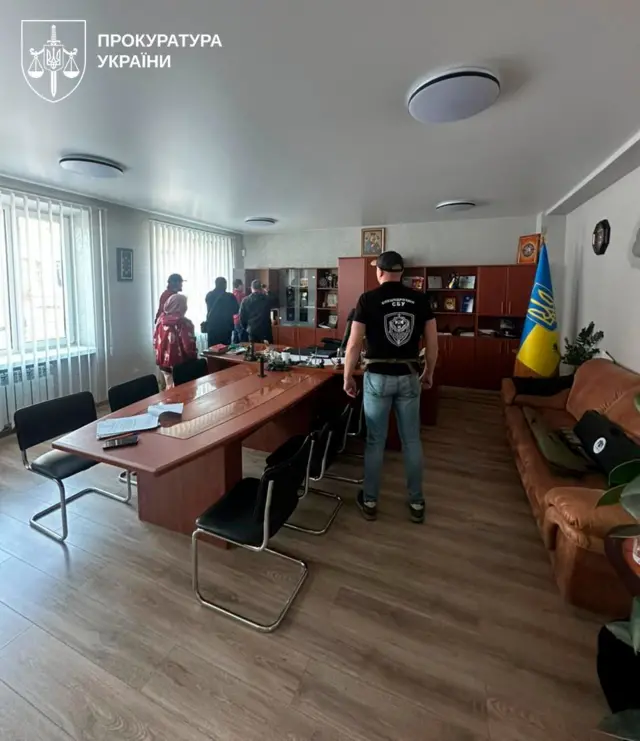 Приміщення, схоже на кабінет начальника поліції, з довгим столом для нарад, стільцями та шафами з документами і нагородами. У центрі стоїть людина у футболці з написом «СБУ», ще кілька людей – біля вікна. Праворуч видно український прапор і диван, над шафою висить ікона Марії з Іісусом.