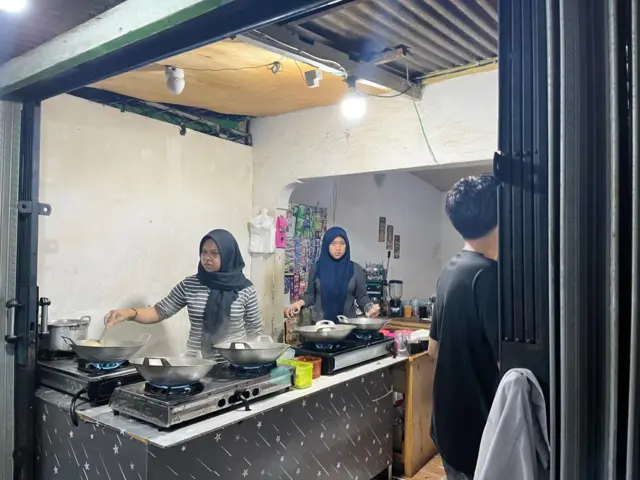 Seorang perempuan berhijab sedang memasak seblak di atas kompor yang menyala dan mengeluarkan asap putih.