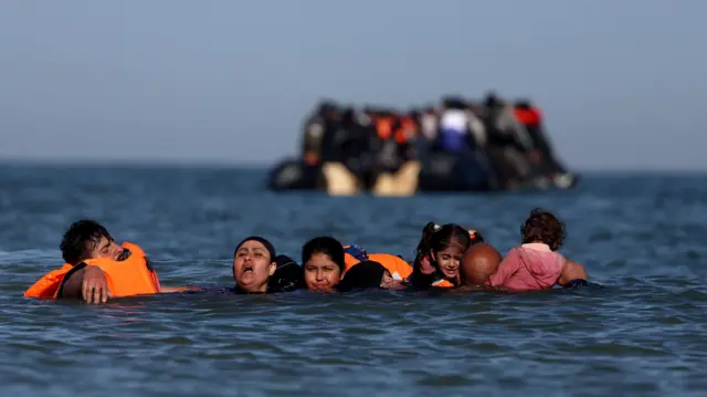 Группа мужчин, женщин и детей в воде на фоне уплывающей в Британию резиновой лодки