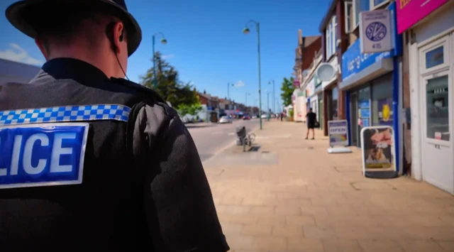 Um policial uniformizado caminha pela calçada da rua, repleta de lojas, sob um céu azul.