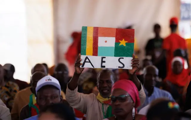 Un homme soulève un tableau sur lequel est écrit "AES" avec les drapeaux des trois pays (Mali, Burkina Faso et Niger).