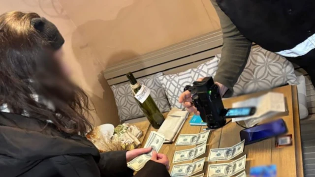 На кадре оперативной съемки слева женщина с заблюренным лицом в черной куртке пересчитывает лежащие на столе долларовые купюры. На столе в центре кроме денег два телефона и пустая бутылка. Справа рука человека с видеокамерой