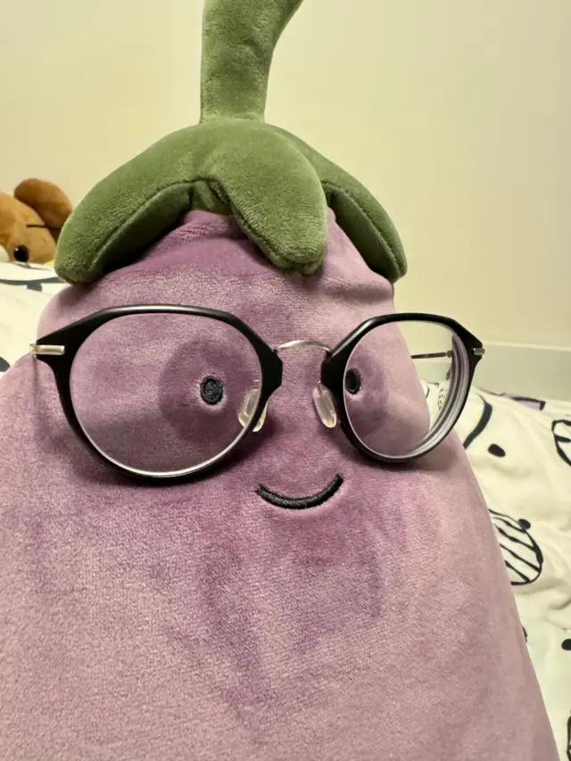 一隻Jellycat出品的紫色茄子毛絨玩具,眼睛下方畫著黑眼圈,頭上戴著一副眼鏡。