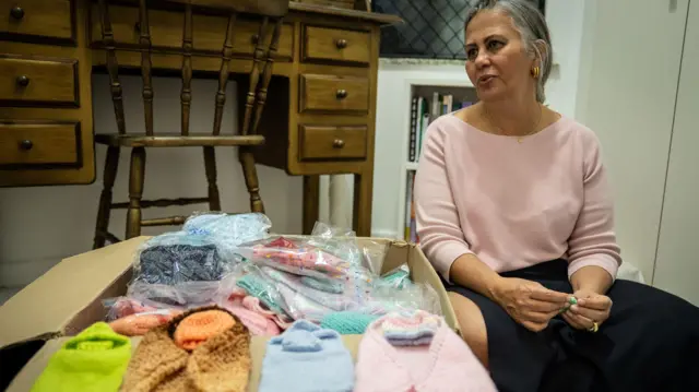 Foto mostra uma caixa de papelão cheia de pequenas roupinhas de tricô e a médica neonatologista Lilia Embiruçu sentada ao lado. 