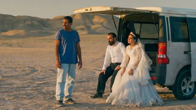 عروس وعريس يجلسان في الجزء الخلفي المفتوح من السيارة، بينما يقف رجل آخر بجانبهما في الصحراء الإيرانية في لقطة من فيلم "كان مجرد حادث".