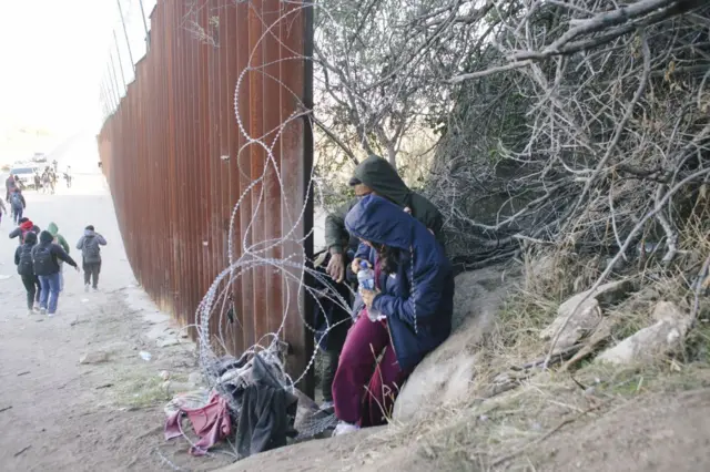 Migrantes passando por uma espaço no muro da fronteira entre Baja California, México, e Califórnia, Estados Unidos