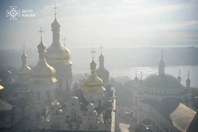 Золоті куполи Києво-Печерської лаври піднімаються над Дніпром, частково затягнуті густим димом і серпанком. Світло пробивається крізь дим, через нього позаду лаври ледь проглядається міський горизонт.