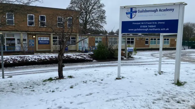El exterior de una escuela, con nieve en el suelo. Hay un edificio de ladrillo, con un gran cartel a la derecha, árboles y una carretera, también con nieve en el suelo.
