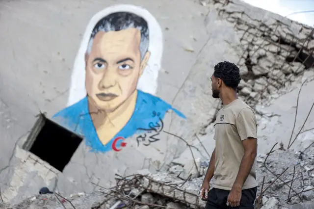 Filistinliler, 27 Haziran 2026'da Gazze Şeridi'nin Gazze kentinde İsrail saldırıları sonucu yıkılan İtalyan Kulesi'nin enkazı üzerine Filistinli sanatçı Abu Karsheli tarafından resmedilen Kamal Adwan Hastanesi Müdürü Dr. Hussam Ebu Safiya'nın portresinin önünden geçiyor.