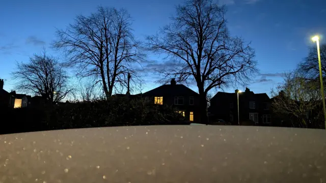 Una capa de nieve cubre el techo de un coche, pero parece que la nieve está en el suelo en lugar del coche. La nieve brilla como si se hubieran esparcido gemas sobre ella, pero son gotas de hielo que reflejan las farolas. El cielo empieza a aclararse y es de un azul intenso. Se ven siluetas de casas detrás del coche, con algunas habitaciones encendidas. A la derecha hay una farola aún encendida y grandes árboles sin hojas.