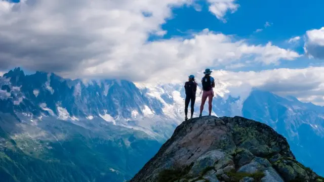 Dua pejalan kaki wanita muda berdiri di atas batu dekat La Flégère, melihat pegunungan Mont Blanc