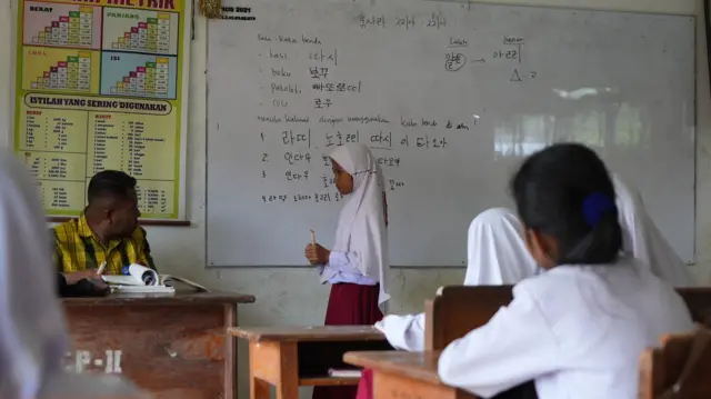 Siswa di SDN Karya Baru berdiri di depan papan tulis yang bertuliskan huruf Hangeul. Siswa itu baru saja mengisi soal yang ditanyakan oleh gurunya.