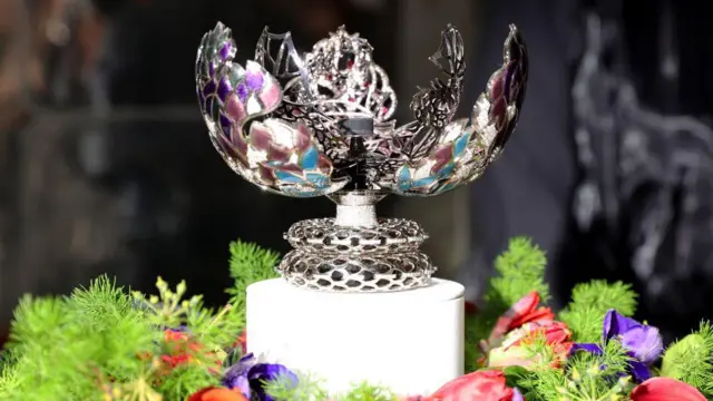 A Game of Thrones-themed egg stands on a white pedestal, surrounded by flowers. Different fragments lean backwards, opening up the egg. It is decorated with silver, purple, and blue colours.