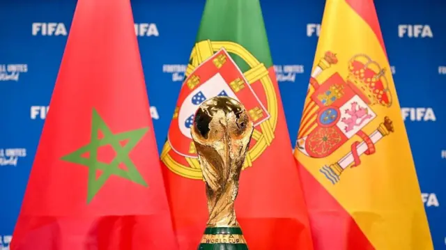 Sur une photo promotionnelle de la Coupe du monde, le trophée doré trône au premier plan, tandis que les drapeaux des trois pays co-organisateurs de l'édition 2030 – le Maroc, le Portugal et l'Espagne – sont disposés de gauche à droite, se chevauchant légèrement à l'arrière-plan devant un panneau bleu sur lequel figure le mot « FIFA » en lettres blanches.