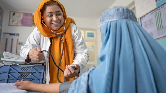 یک داکتر که لباس سفید و چادر زرد روشن دارد در حال معاینه یک ن دیگر که چادری آبی دارد.