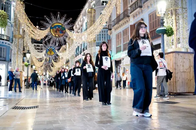 Siyah giyinmiş kadınlar tek sıra halinde ellerinde bir kadın fotoğrafı olan kağıtlarla sokakta yürüyor, sokak ışıltılı süslemelerle kaplı