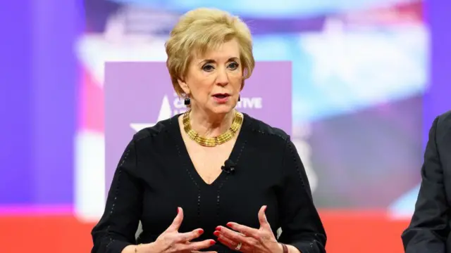 Linda McMahon, với mái tóc ngắn màu vàng, mặc một chiếc váy đen và đeo một sợi dây chuyền vàng dày, dùng cả hai tay để diễn tả trong khi ngồi tại một buổi thảo luận tại Hội nghị Hành động Chính trị Bảo thủ (CPAC).