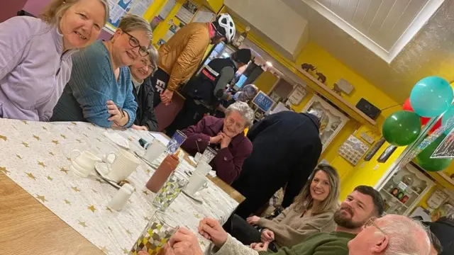 Un grupo de siete personas se sienta alrededor de una mesa en un café. Margaret preside la mesa, con un abrigo morado y sonriendo. Todos miran a la cámara y sonríen. A la derecha, sobre sus cabezas, cuelgan globos del techo.
