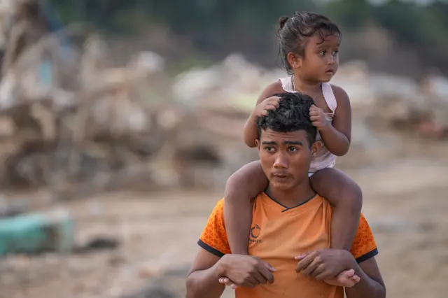 Warga menggendong anaknya di tengah pusat Kota Kuala Simpang yang luluh lantak akibat banjir bandang di Aceh Tamiang, Aceh, Sabtu (6/12/2025).