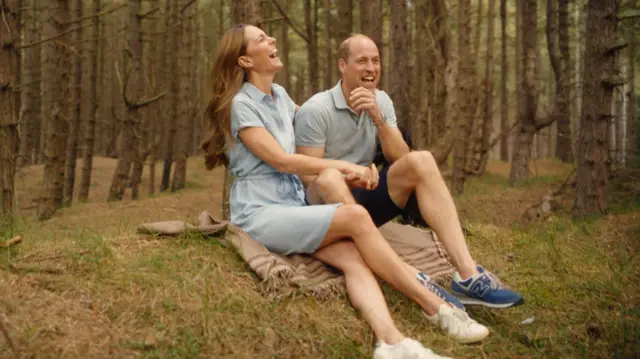 Kate e William rindo sentados na grama, em meio a árvores, em cena do vídeo filmado em Norfolk.