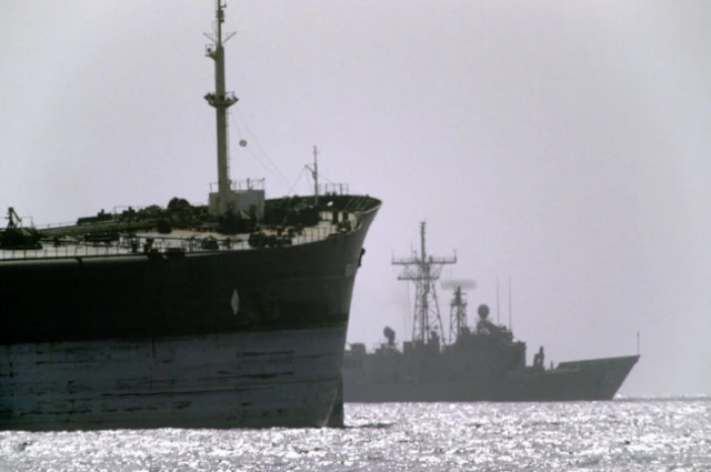 A US warship in the background travels alongside a cargo ship during Operation Earnest Will in the Strait of Hormuz.