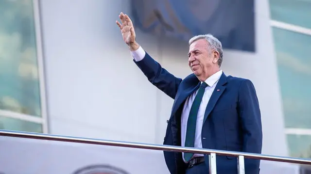 Mansur Yavaş bir seçim otobüsü üzerinde mavi ceketiyle sağ elini kaldırarak halkı selamlıyor.
