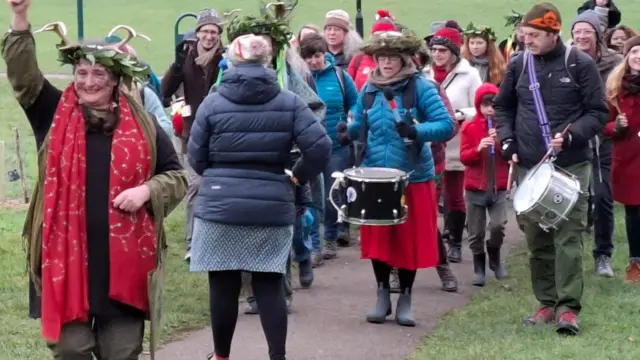 Una procesión de adultos y algunos niños camina por el parque hacia la cámara. Llevan ropa de abrigo invernal. La procesión la encabeza Konni, quien viste ropa verde caqui y una guirnalda de hojas verdes en la cabeza, con algunas marcas verde oscuro pintadas en la cara. Algunas personas tocan tambores y un niño toca la flauta dulce.
