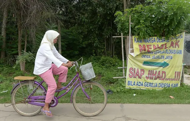 Ilustrasi: Seorang warga bersepeda melewati spanduk bertuliskan "Kami Warga Desa menolak menolak pembangunan gereja. Siap Jihad bila ada gereja di desa kami"