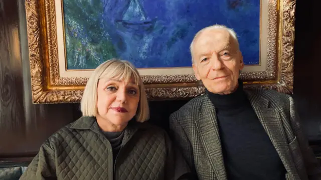 Una mujer madura con pelo claro y un hombre maduro de pelo canoso vestidos causual pero elegantemente y sentados frente a un cuadro de Chagall