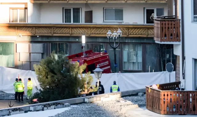 Bir binanın dışında beyaz bir branda var. Aynı zamanda yeşil üniforma giymiş güvenlik görevlileri ve kırmızı bir araç var