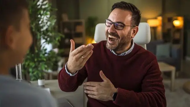 Молодий чоловік у светрі і окулярах щось жваво розповідає іншому чоловіку і посміхається