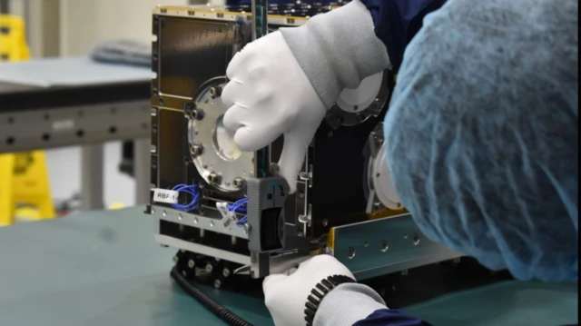 صورة نشرتها وكالة الفضاء السعودية لما قالت إنهم مهندسون سعوديون يرتدون قفازات وأفنعة وجه وأغطية رأس يعملون على القمر الصناعي شمس بجوار علم المملكة الأخضر