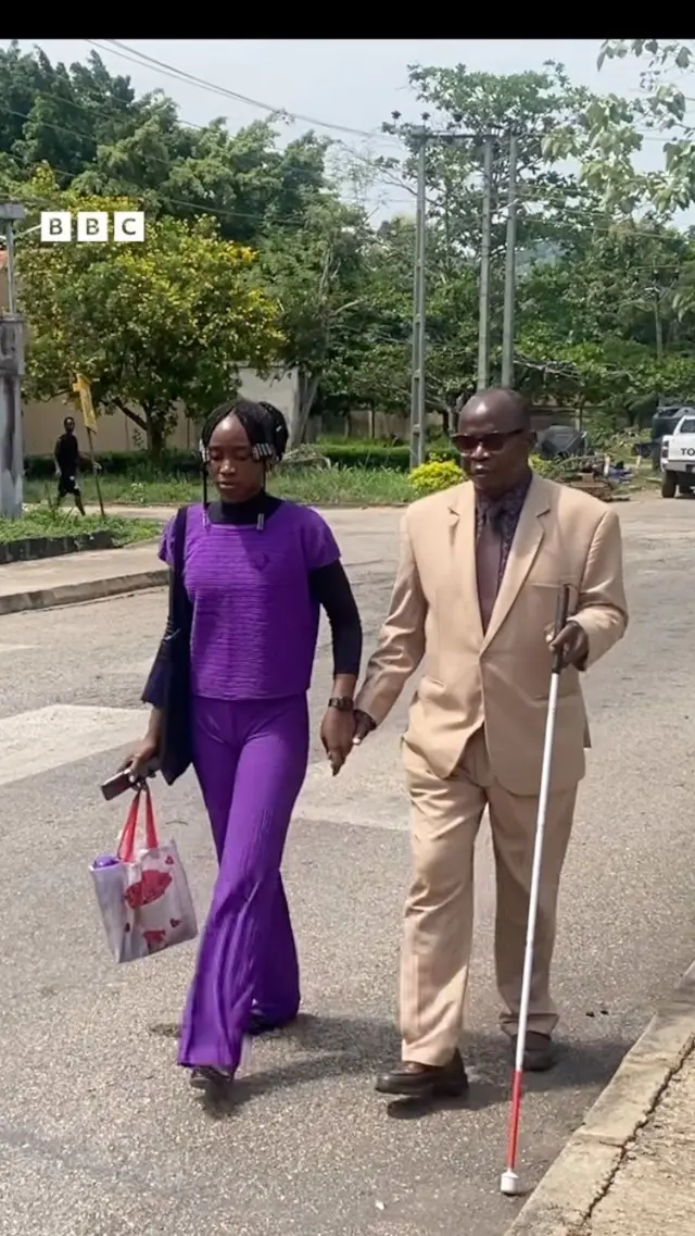 Emmanuel Modurotimi Olubodede to wọ asọ kootu alawọ paali, mu ọpa kan lọwọ lati rin, nigba ti obinrin kan to wọ asọ sokoto ati bilaosi alawọ pọpu fa lọwọ lasiko to n rin