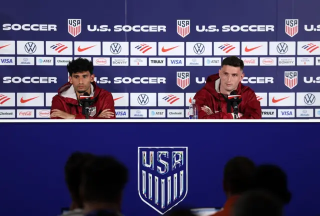 Ricardo Pepi y Matthew Freese con chaquetas rojas del uniforme de la selección de EE.UU. Ambos están sentados y tienen micrófonos al frente. Detrás tienen una pared llena de símbolos de marcas y escudos de USA y las palabras U.S. Soccer. Al frente hay periodistas