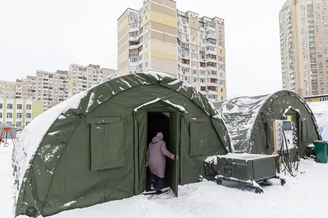 Польові намети зеленого кольору, вкриті снігом, встановлені серед багатоповерхових житлових будинків. До одного з наметів заходить жінка в зимовому одязі. Поруч стоїть обладнання, схоже на генератори або системи обігріву, а навколо — засніжений двір.
