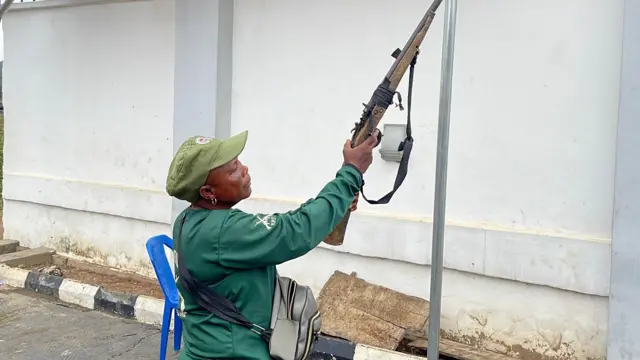 Mama Marian Alarape Iyanuore ree to wọ asọ ologun alawọ ewe, o kọ ẹyin si ile kan, to si duro, na ibọn sakabula to wa lọwọ rẹ soke bii pe o fẹ yin ibọn.