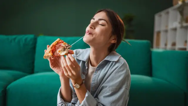 Kobieta delektuje się kawałkiem pizzy, ma na sobie niebieską koszulę, w tle widać zieloną sofę 