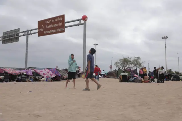 Niños jugando con una pelota en un área de arena junto a carpas, con un letrero fronterizo de Chile al fondo.