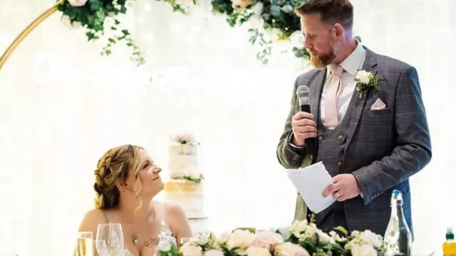 Dave, vestidocódigo promocional da betânianoivo, discursa olhando para Charlotte durante o casamento, com o bolo ao fundo