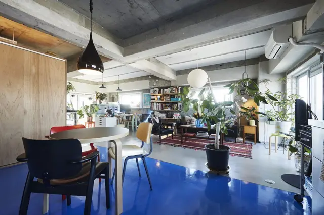 Espacio interior de concepto abierto con suelo azul brillante, mobiliario moderno, plantas de interior y vigas de hormigón visto.