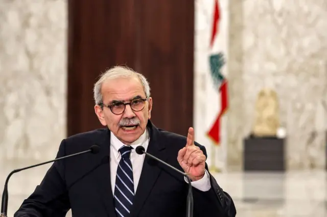  رئيس الحكومة اللبنانية، نواف سلام واقفاً وأمامه ميكرفون، مرتدياً سترة سوداء وقميصاً أبيض وربطة عنق يمتزج لونها ما بين الأبيض والأسود.