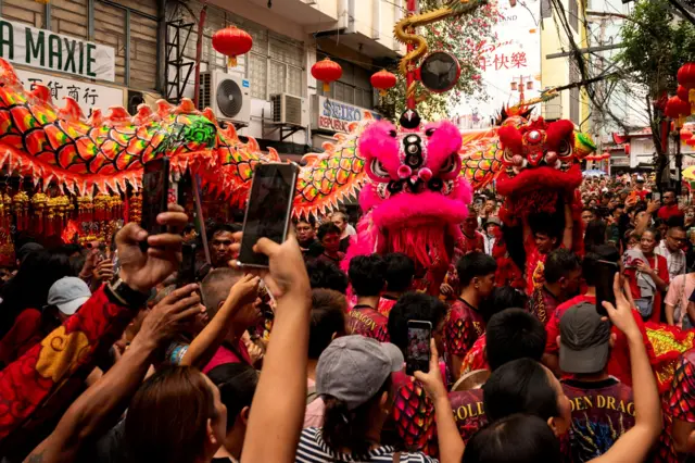 Толпа людей (многие снимают на телефон) смотрит на фигуры льва и дракона, в которых находятся танцоры.