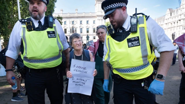 Gözaltına alınan ileri yaştaki kadının elinde, "Soykırıma karşı çıkıyorum, Palestine Action'ı destekliyorum" yazıyor. Yanında iki üniformalı polis var.