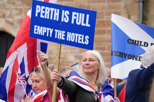 یک زن در پرت در اسکاتلند به نشانه اعتراض به اسکان پناهجویان در هتل‌ها پلاکاردی در دست دارد که بر روی آن نوشته شده: «پرت پر است، هتل‌ها را خالی کنید»