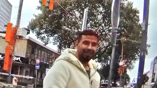 A dark-haired man with a beard and white fleece jacket smiles unsuspectingly at our reporter. He is standing outdoors with a parade of shops, a tree and street furniture surrounding him. 