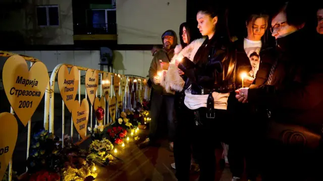 Неколико људи пали цвеће код металне ограде на којој су бела срца са именима жртава у Кочанима